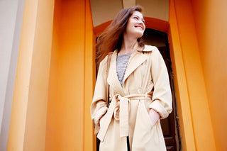 Souriante jeune femme portant un trench beige classique, prête à sortir dans une rue vivante.