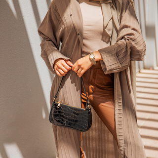 Une femme à la mode avec une montre dorée tenant un sac à main en cuir noir texturé, vêtue d'une tenue plissée dans les tons brun et beige.