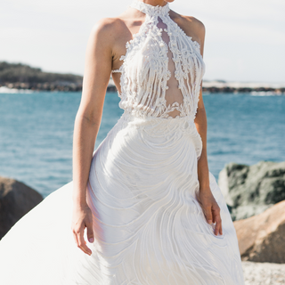 Une mariée portant une robe de mariée dos nu à détails en dentelle, se tenant sur la plage avec la mer en arrière-plan.