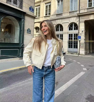 Jeune femme souriante portant une veste aviateur en cuir beige, un t-shirt blanc et un jean bleu dans une rue de la ville.