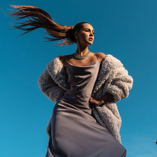 Portrait dynamique d'une femme élégante vêtue d'une robe satinée grise avec un manteau en peluche sur les épaules, ses cheveux au vent, incarnant l'esprit de s'exprimer librement à travers un style personnel et distinctif.